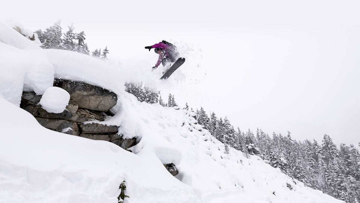 Skier in purple jacket airs off snowy rock cliff, spraying powder