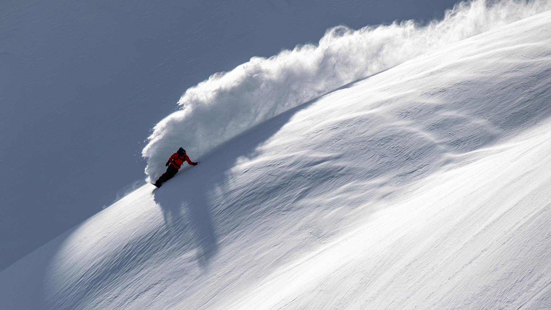 Snowboarder in red jacket carving deep powder, spraying large snow wave