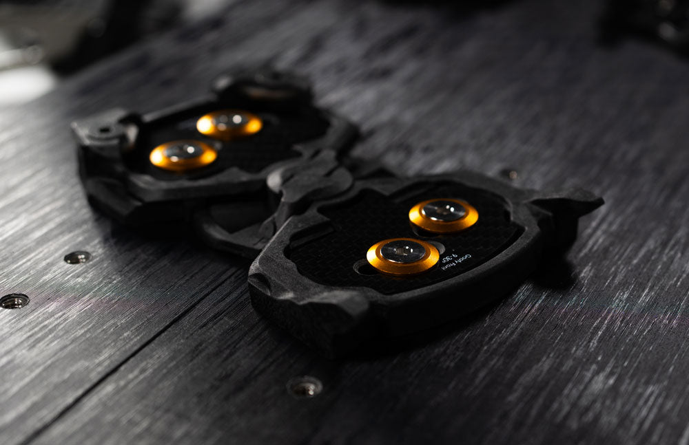 Black carbon fiber bike cleats with orange bolts on wooden surface