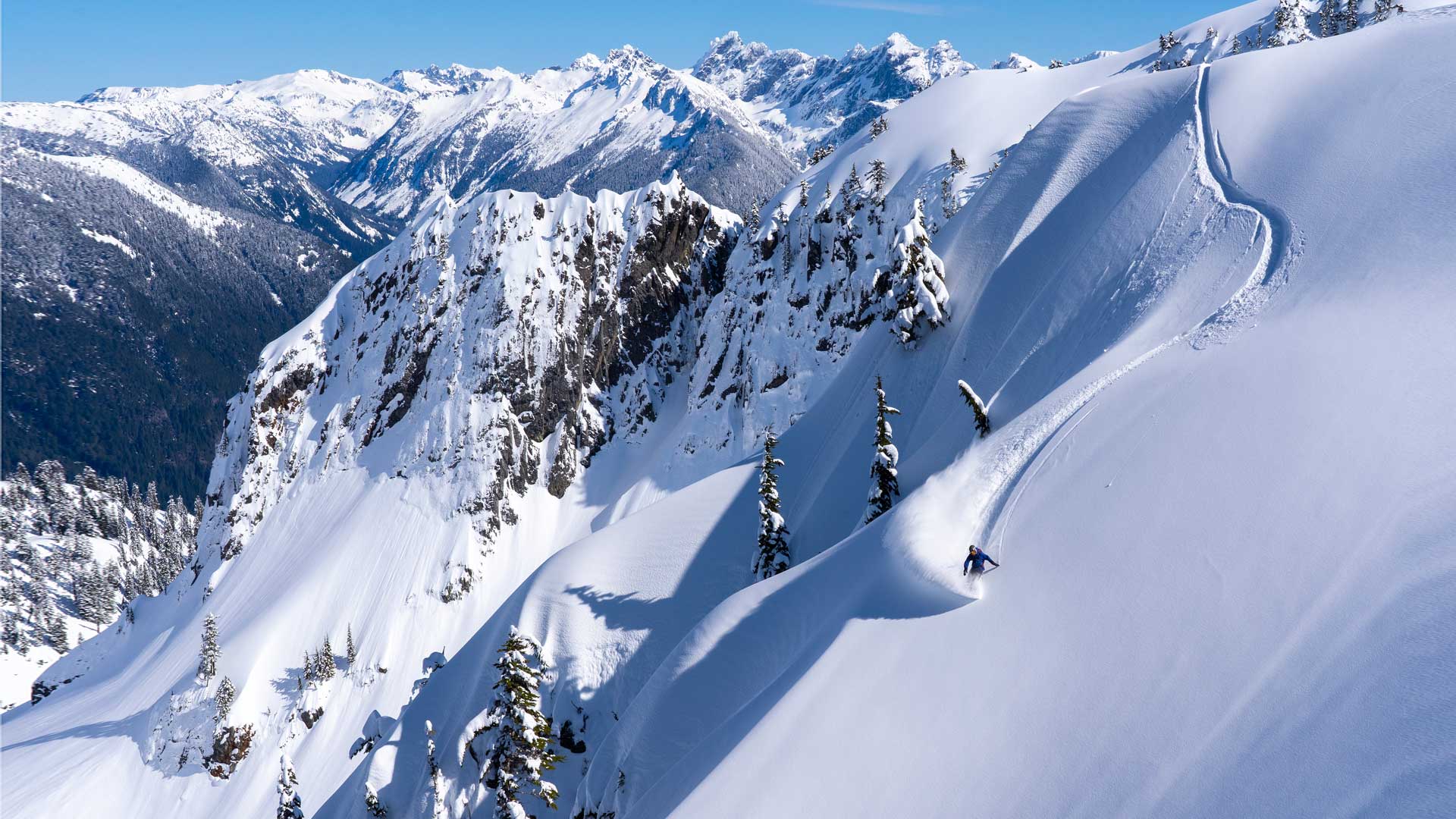 Skier descending steep powder slope with snowy mountain range backdrop