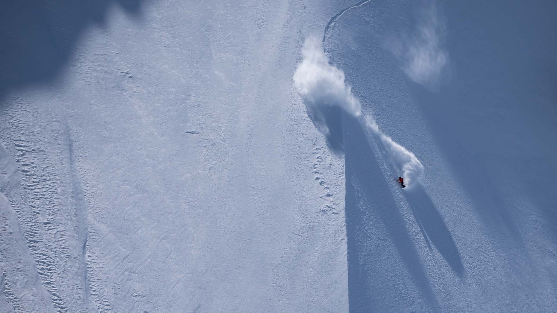 Skier in red jacket descends steep snowy slope, kicking up powder spray