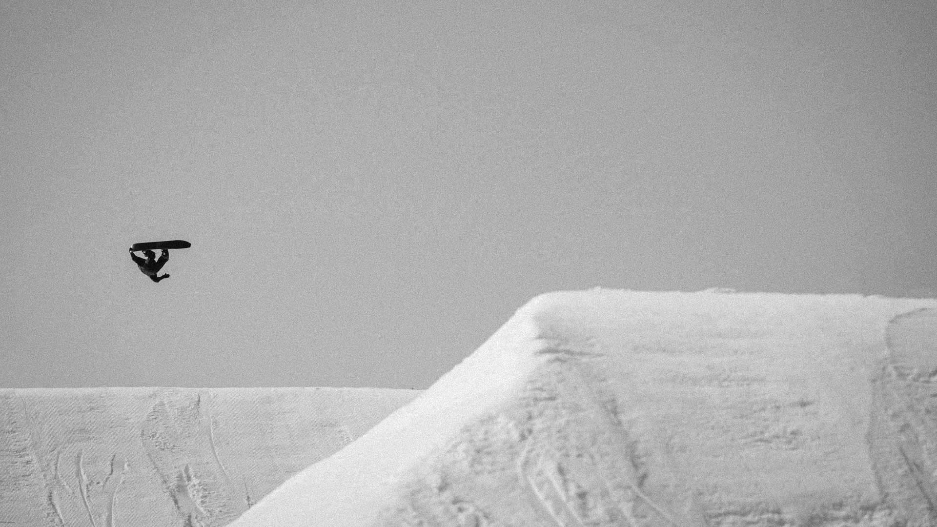 Snowboarder grabbing board mid-air above snowy jump, black and white