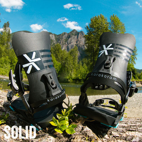 Black snowboard bindings on riverbank rocks with mountain backdrop