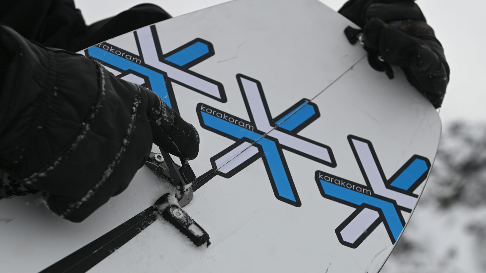 Gloved hands adjusting splitboard binding on white snowboard with Karakoram logos