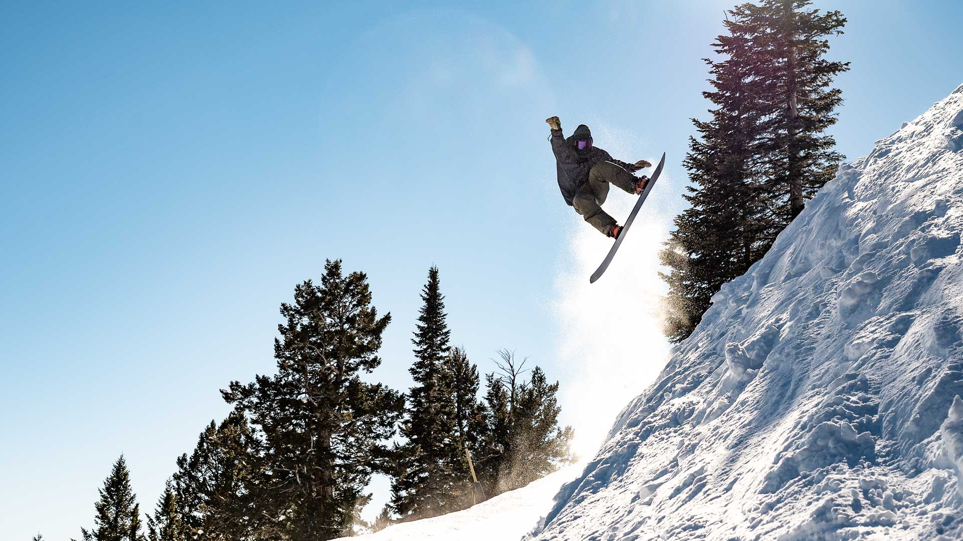 Snowboarder jumping off snowy cliff against blue sky with pine trees