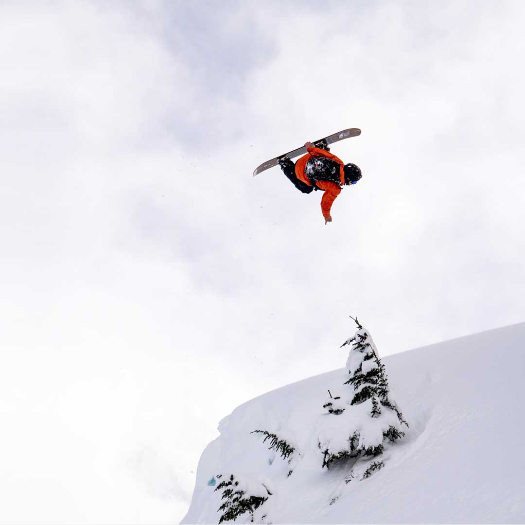 Snowboarder in orange jacket grabs board mid-air above snowy cliff