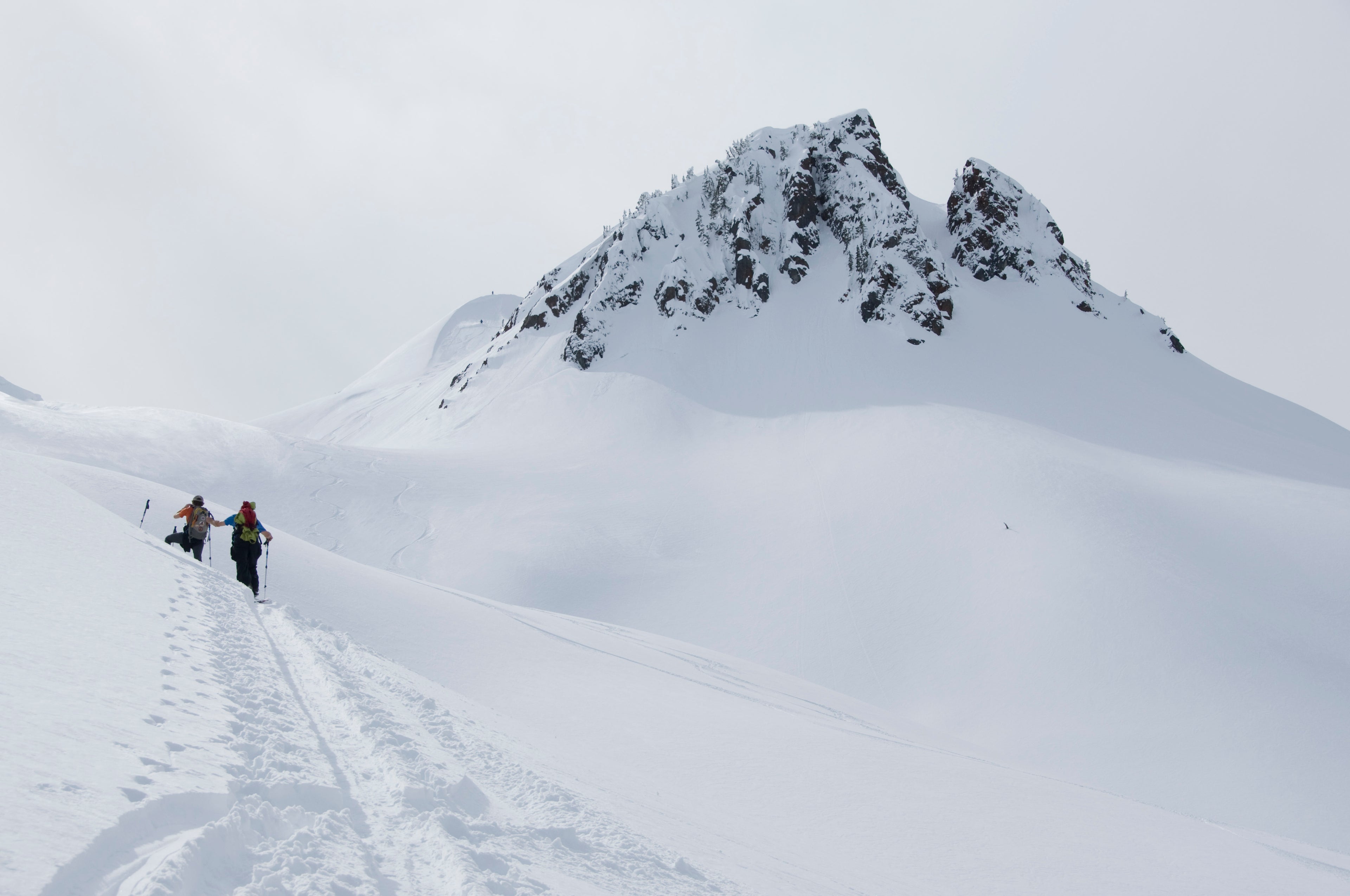 Two backcountry skiers climbing snowy ridge below rocky mountain peak